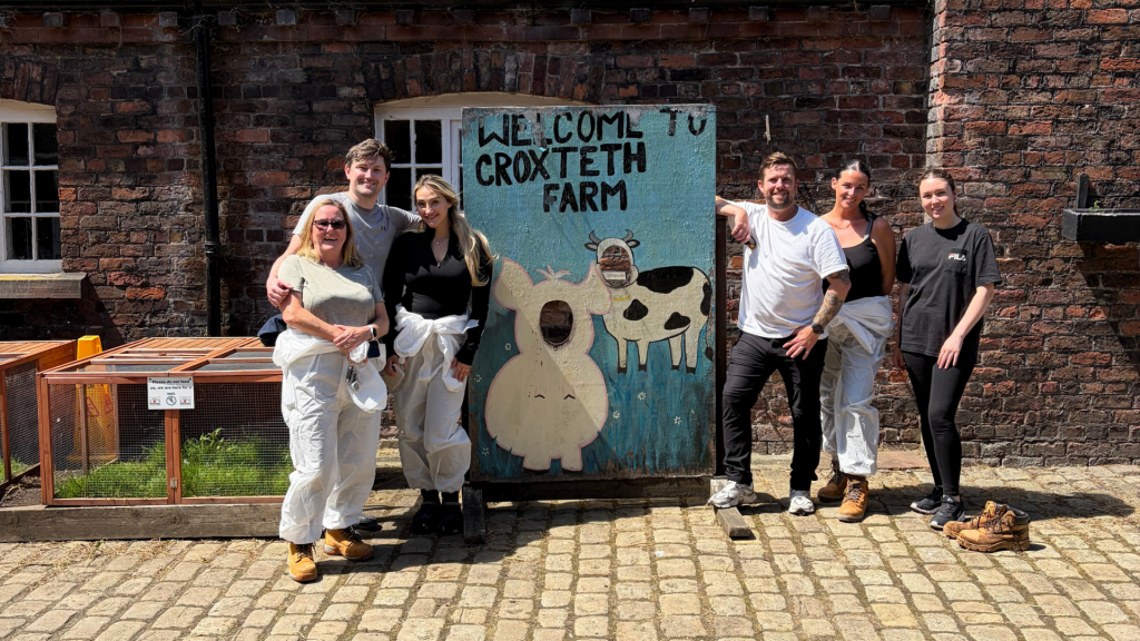 Six people stood next to a sign saying 'Welcome to Croxteth Park Farm'. They're smiling with their arms around each other. It's a bright and sunny day.