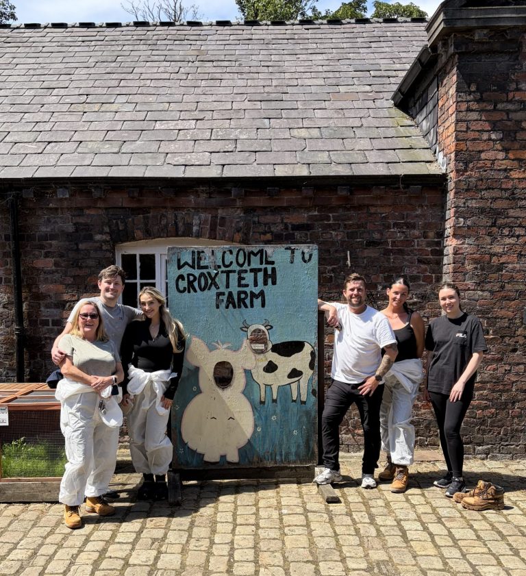 Six people stood next to a sign saying 'Welcome to Croxteth Park Farm'. They're smiling with their arms around each other. It's a bright and sunny day.