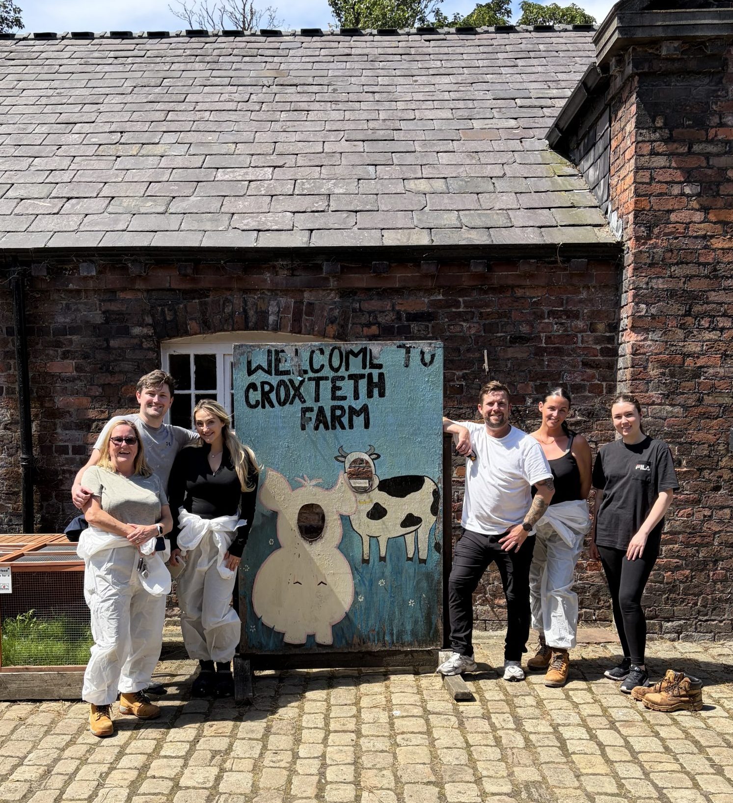 Six people stood next to a sign saying 'Welcome to Croxteth Park Farm'. They're smiling with their arms around each other. It's a bright and sunny day.
