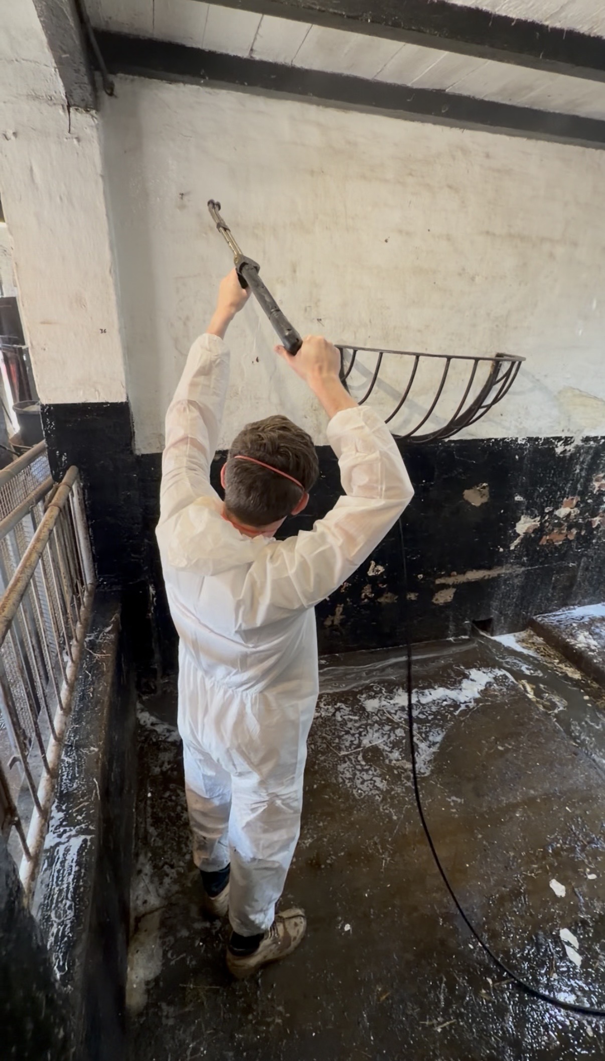 A man with his back to the camera. He's using a jet wash to clean a wall. He's wearing a while overall and a mask.
