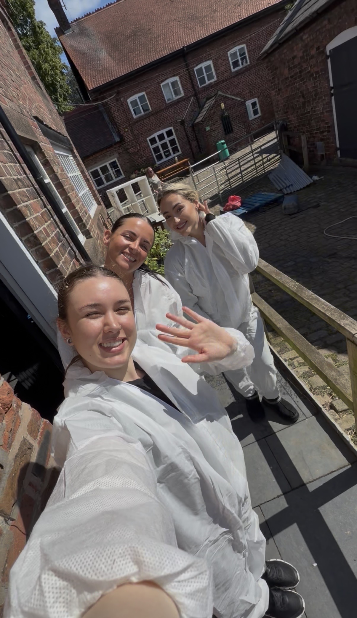 Three women in white overalls smiling at the camera. One is waving at the camera. The sun is shining on them.