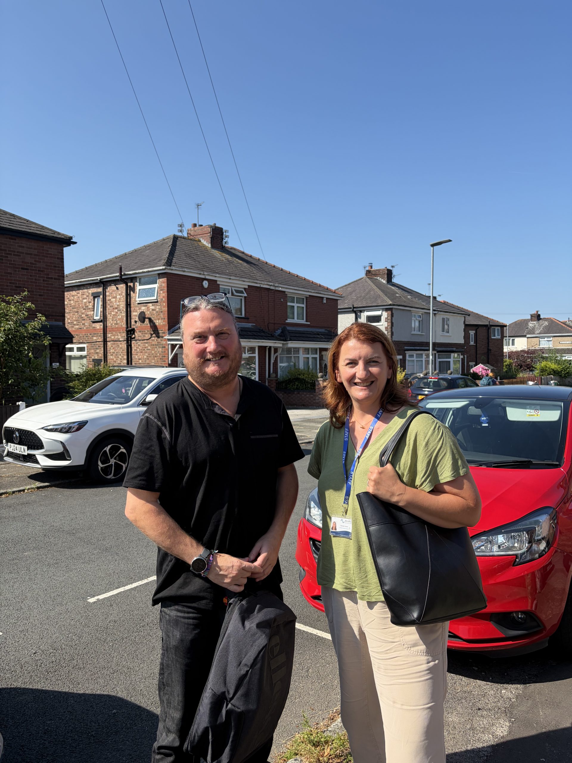 A man and a woman stood next to each other in a street. Houses and cars can be seen the the background. The sky is clear and blue.