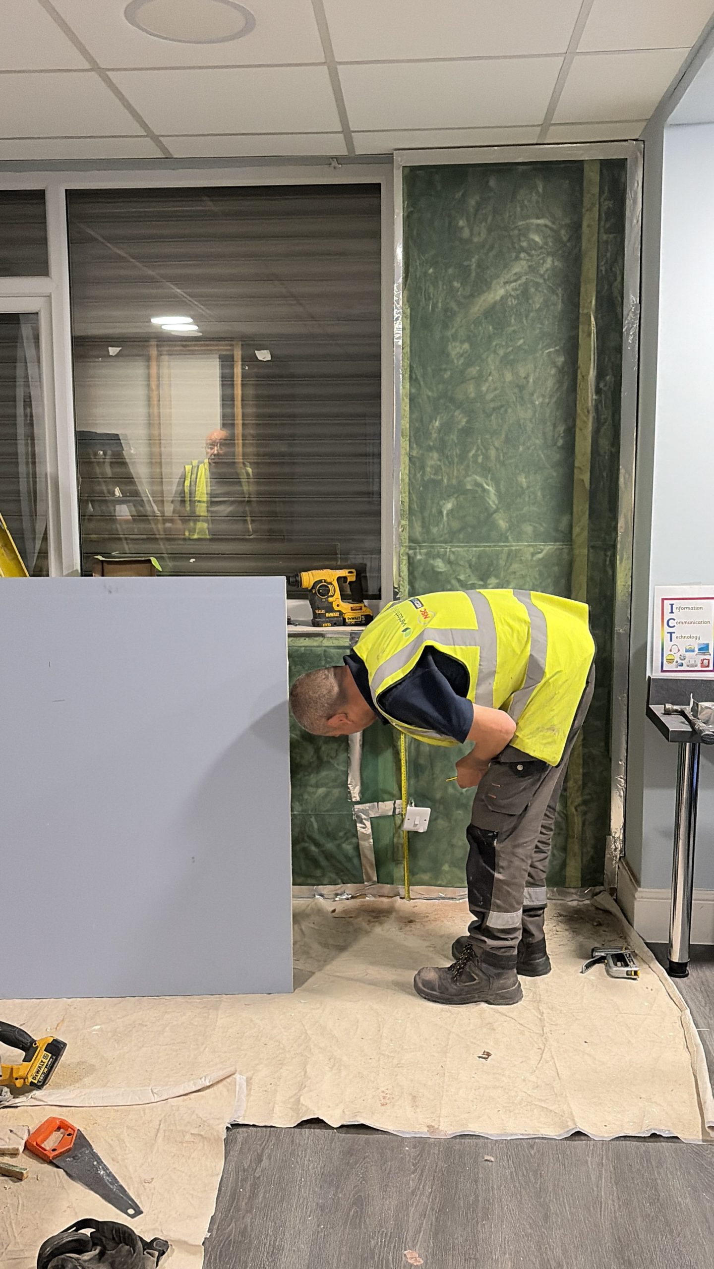A man bending down to measure part of a wall. The wall has freshly installed insulation with a protective cover on it.