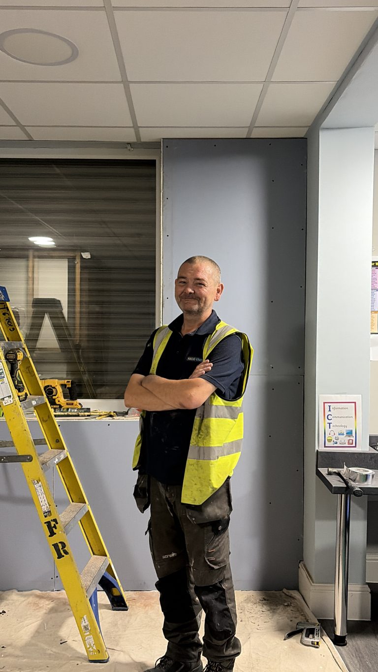 A man stood side-on with his arms crossed, smiling at the camera. He's wearing a high-vis vest, and there is a ladder next to him.